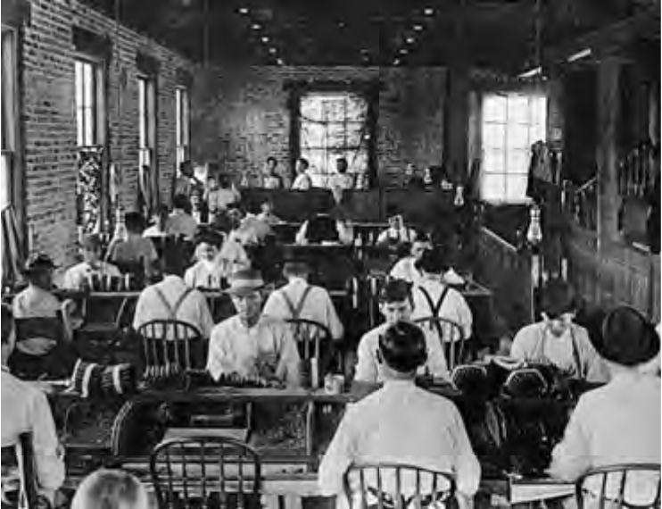 A historical black and white photo of a classroom filled with students at desks, engaged in work, with brick walls and large windows.
