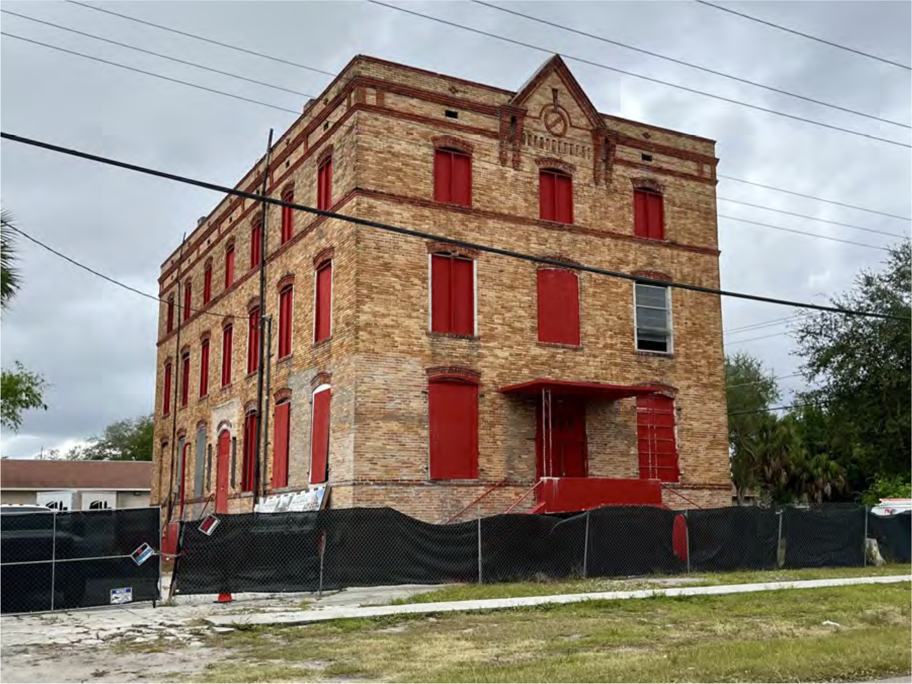 Abandoned brick building with red windows and doors, surrounded by a fence, under a cloudy sky.
