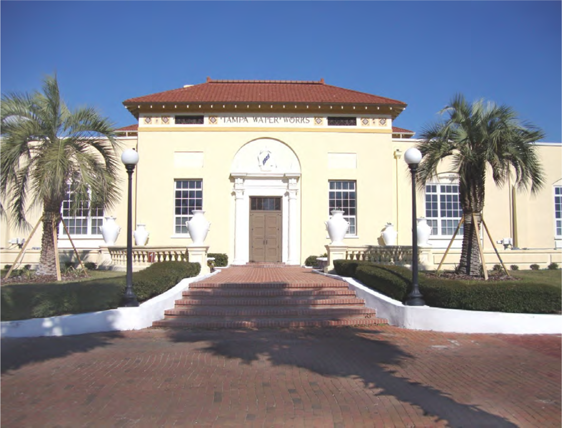 Historic Tampa Water Works building with palm trees, white urns, and red brick stairs against a clear blue sky.