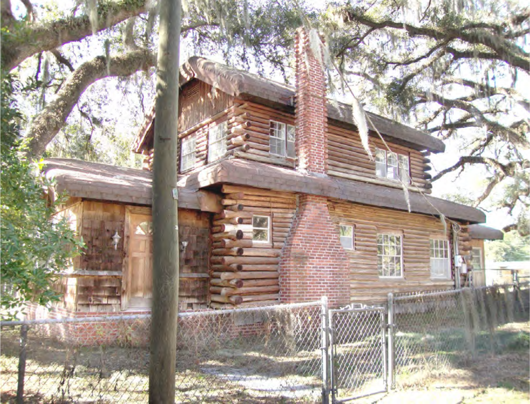 A log cabin with a brick chimney surrounded by trees and a fenced yard.
