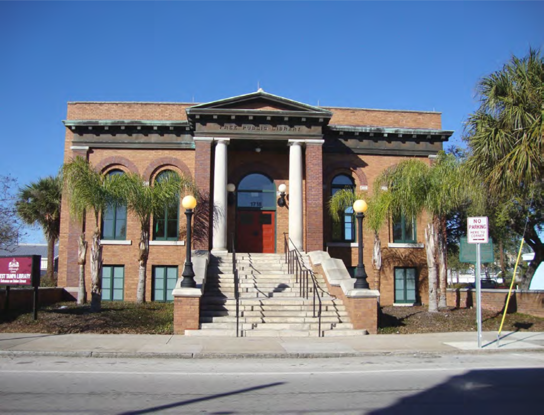 Historic brick building with columns, palm trees, and stairs leading to a red door under a clear blue sky.