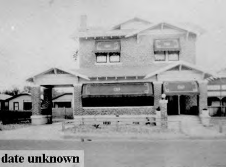 A historical two-story brick house with awnings, featuring a front porch and surrounding landscape; date of the photograph is unknown.