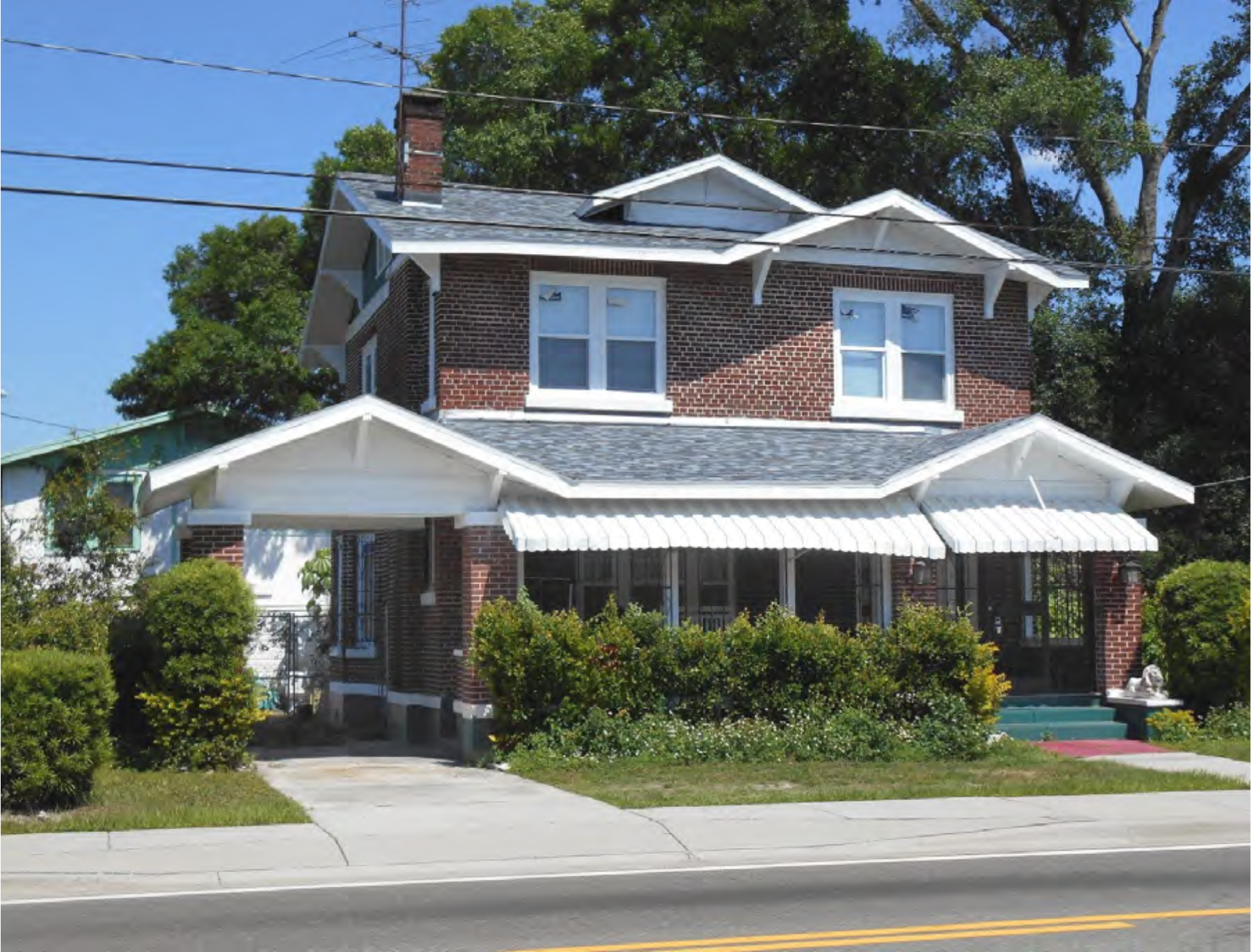 Two-story brick house with a front porch, white awning, and green landscaping on a sunny day.