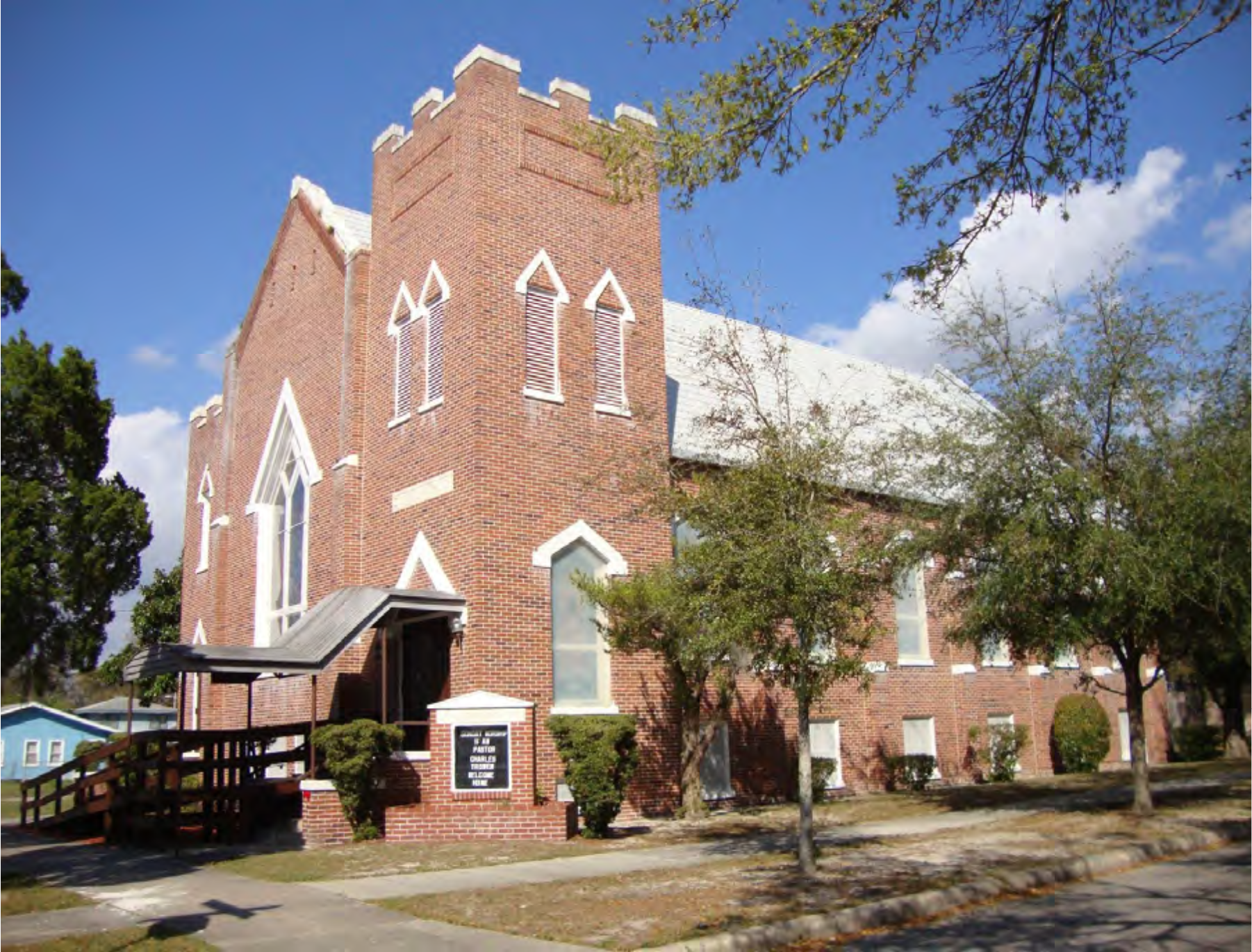 A brick church building with decorative windows, a peaked roof, and trees in front, under a blue sky.