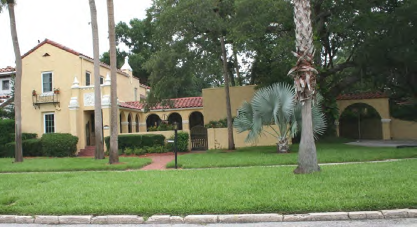 A Spanish-style house with a red tiled roof, palm trees, and lush green landscaping.