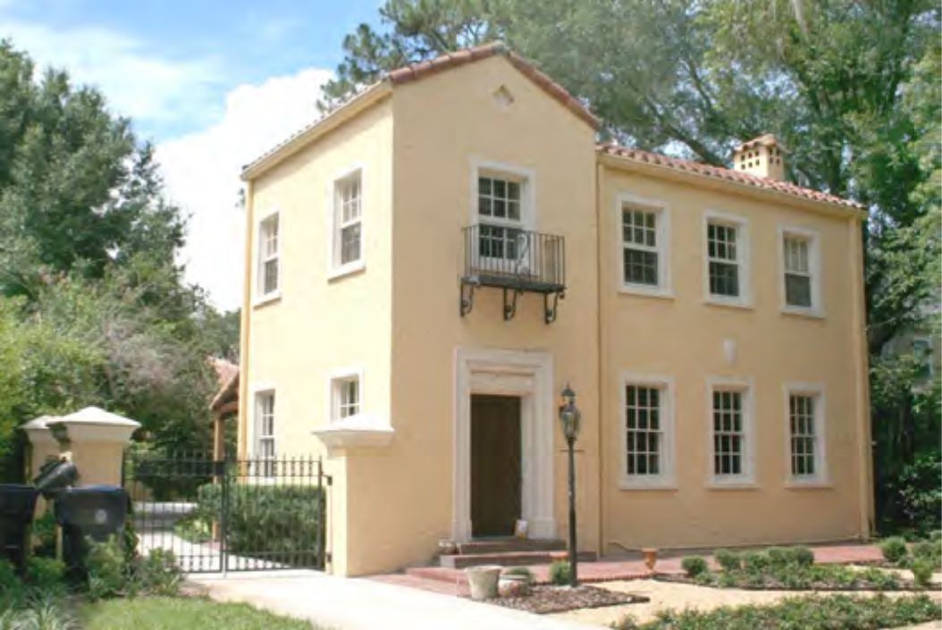 A two-story yellow house with a balcony, surrounded by greenery and a gate, featuring a walkway and lamp post.