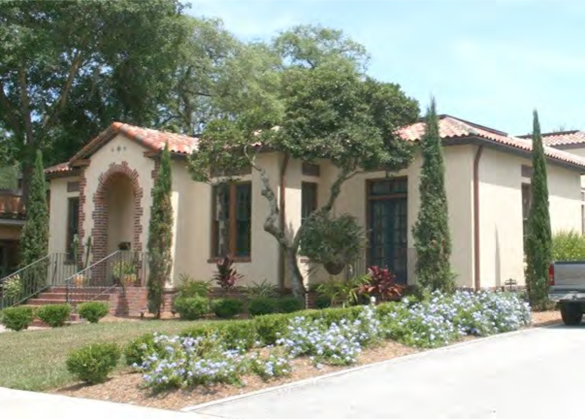 A charming Mediterranean-style house with a curved entryway, surrounded by landscaping, tall trees, and blue flowers under a clear sky.