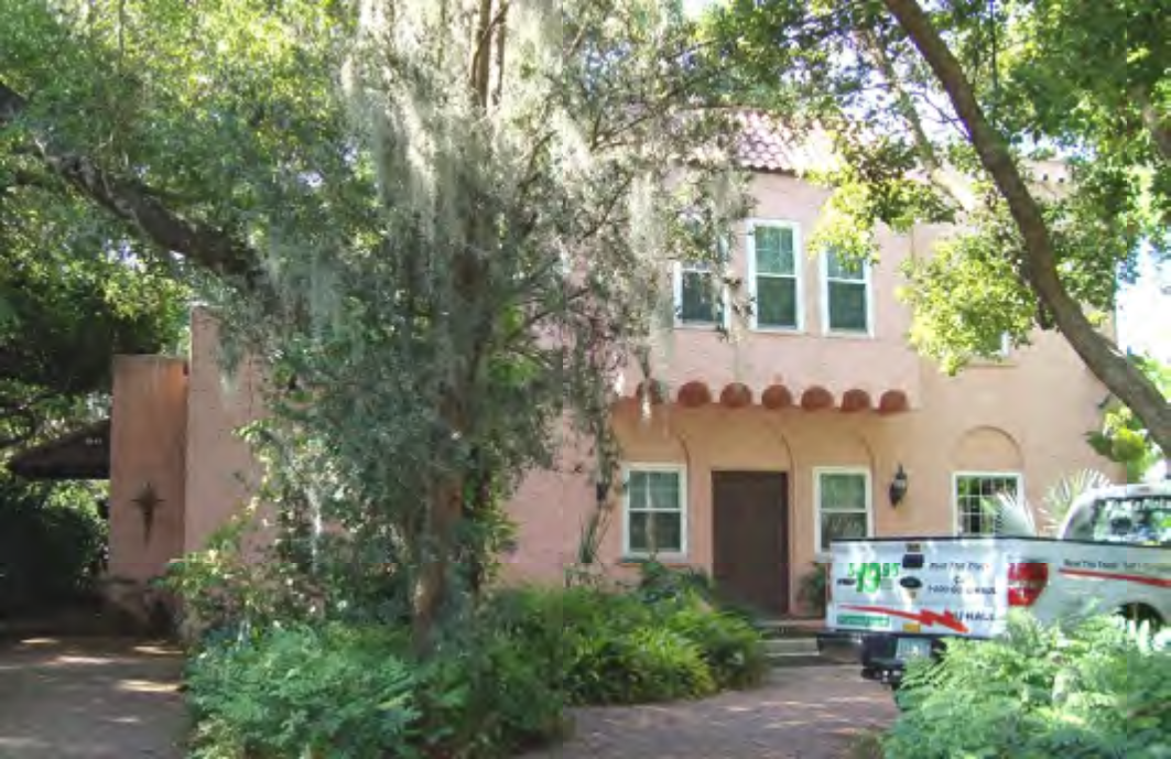 A pink two-story house surrounded by lush greenery and trees, with a white truck parked in front.