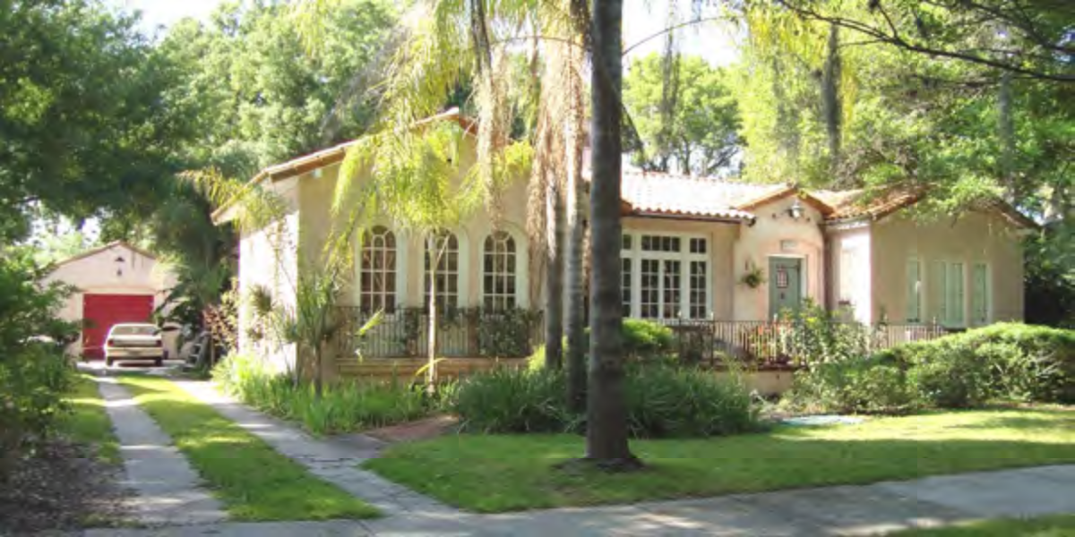 A charming house with a porch, surrounded by greenery and palm trees, located on a sunny street.