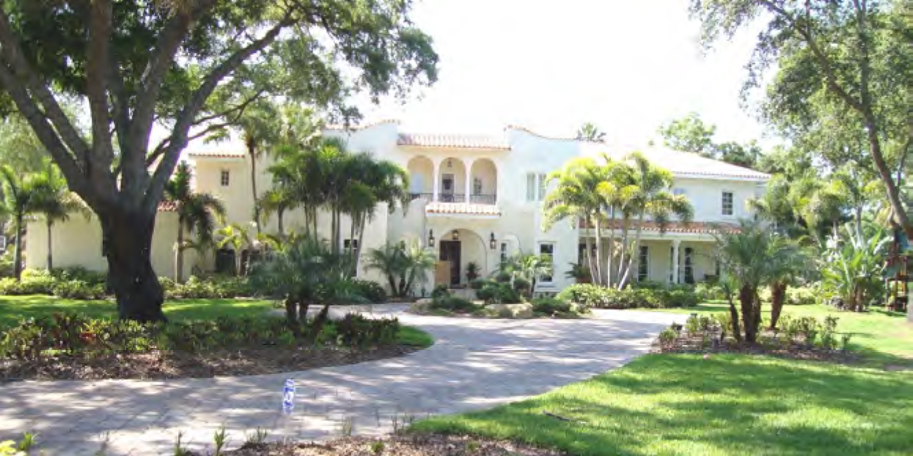 Spacious white mansion surrounded by palm trees and landscaped gardens on a sunny day.