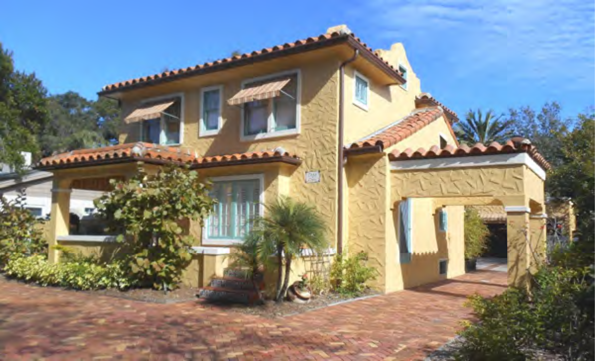A two-story yellow stucco house with a red-tiled roof, surrounded by greenery and a brick-paved driveway.