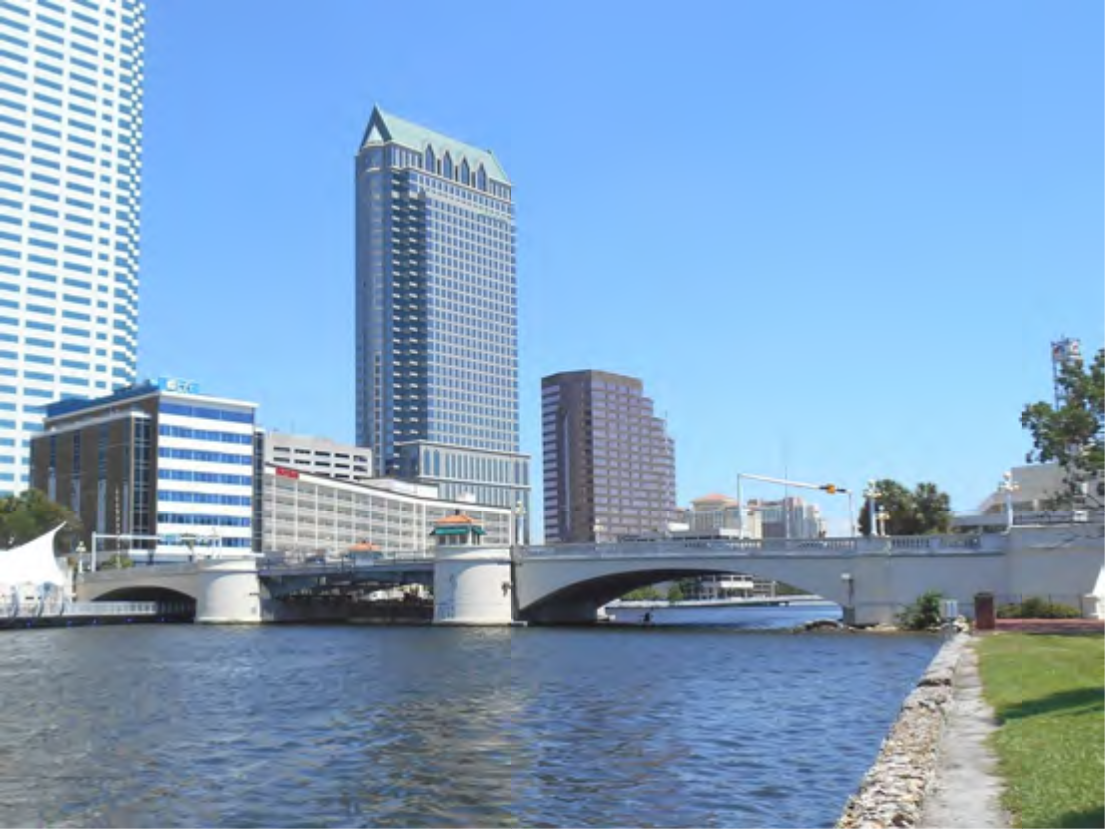 City skyline with modern buildings alongside a river, featuring a bridge and clear blue sky.