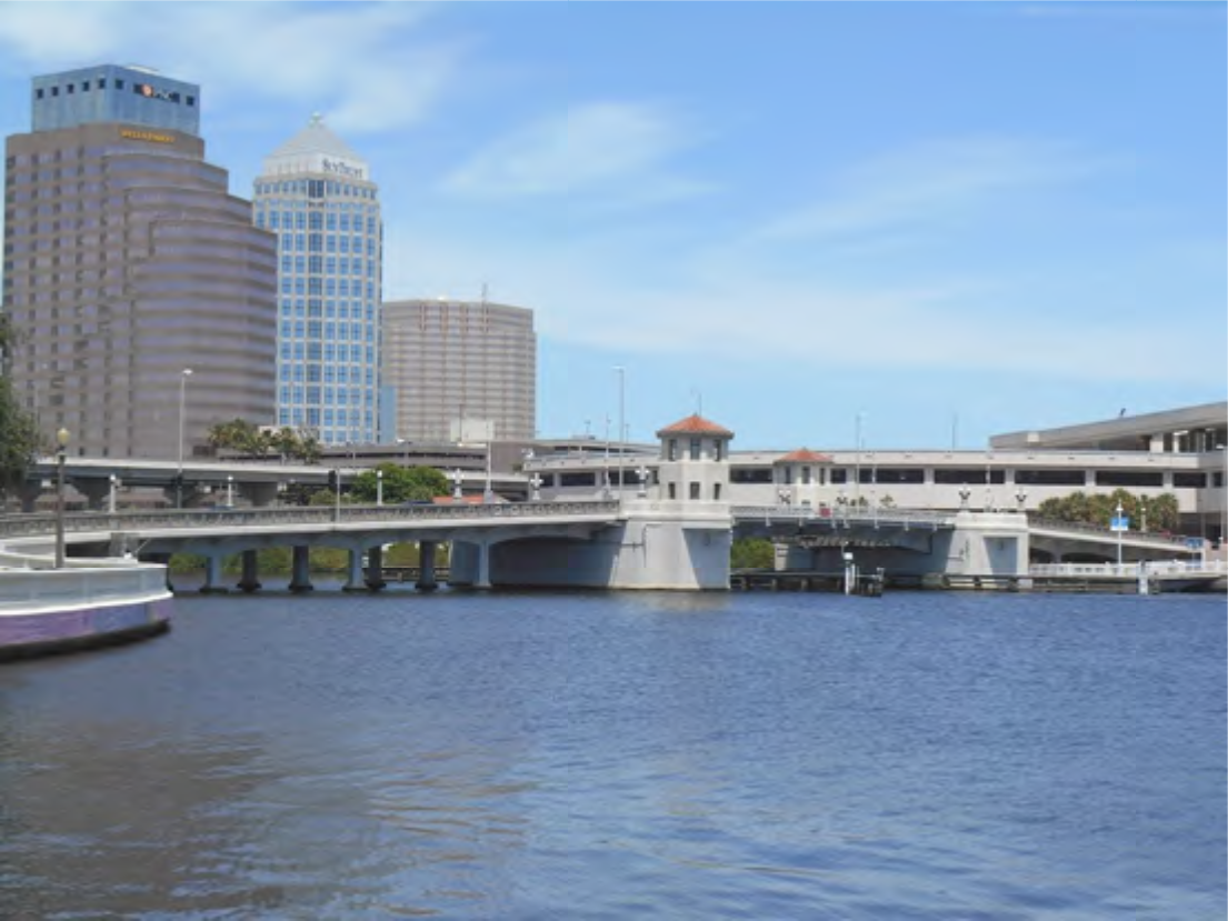 A view of a bridge spanning a river with urban buildings in the background under a clear blue sky.