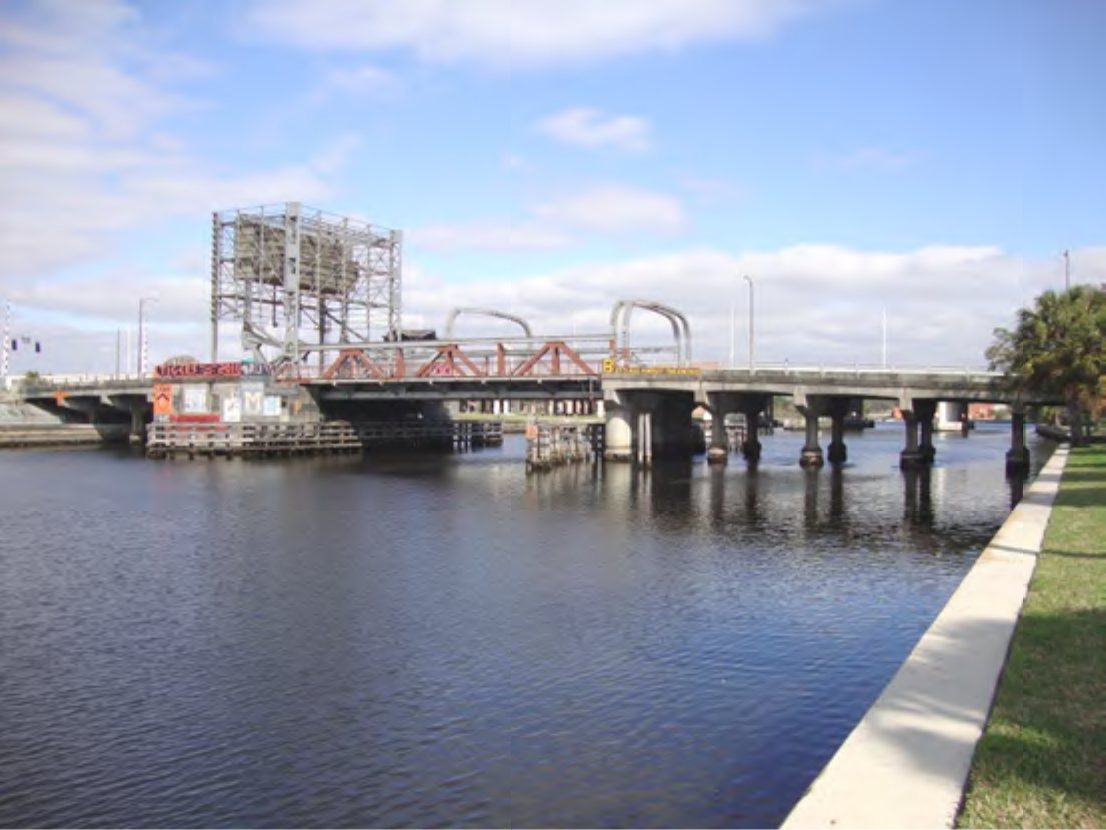 A drawbridge over a calm river with blue skies and scattered clouds, alongside a grassy bank and industrial elements.