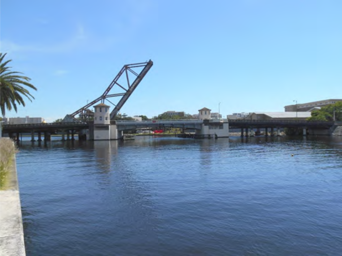 A drawbridge over a calm river with palm trees in the foreground and clear blue sky.