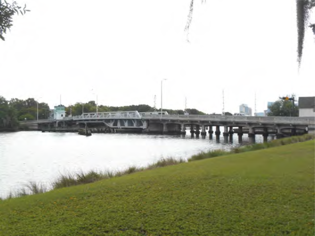 A bridge over a calm waterway, flanked by grassy areas and trees, with buildings in the background under a cloudy sky.