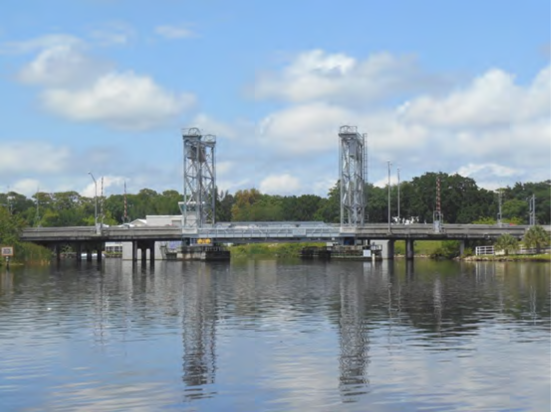 A drawbridge over a calm river with a blue sky and fluffy clouds in the background.