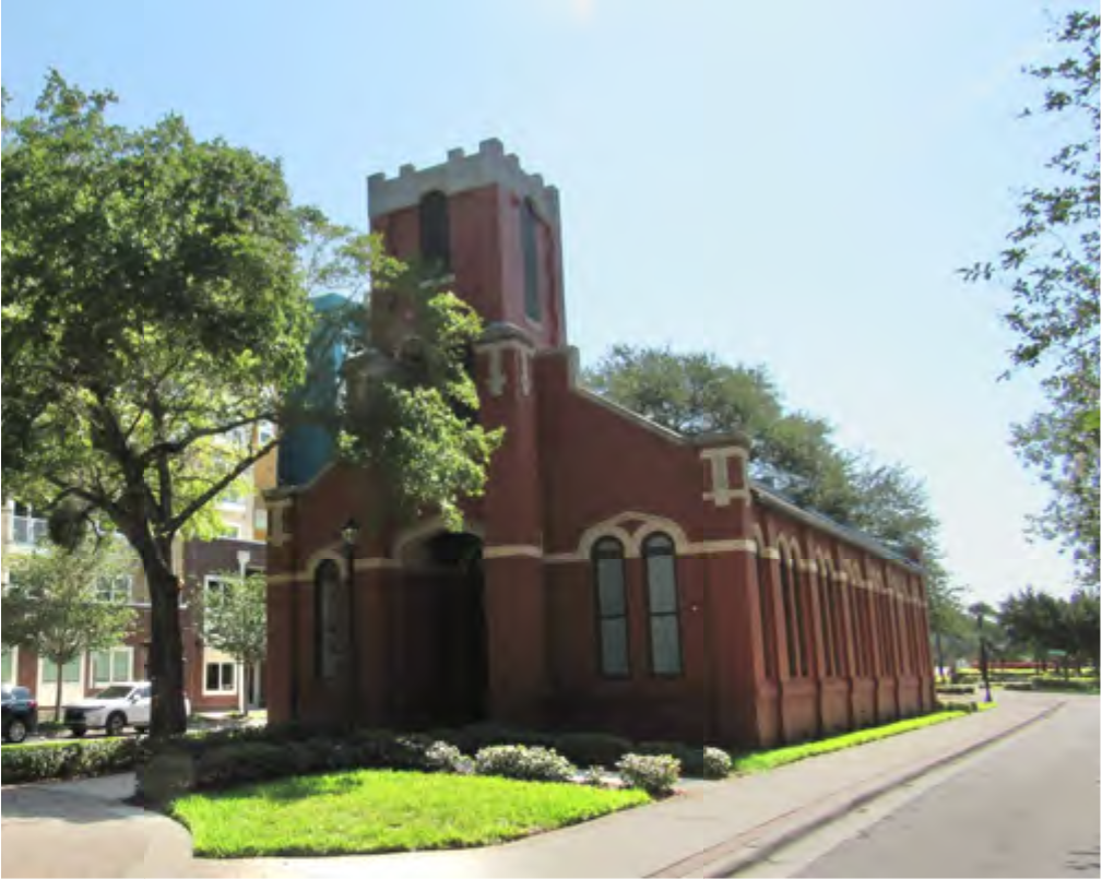A historic red brick church with a tower, surrounded by green grass and trees, set against a clear blue sky.