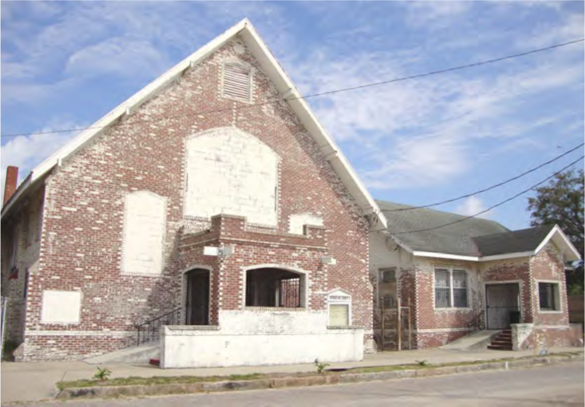 An old brick building with a gabled roof and boarded windows, situated on a street under a blue sky.