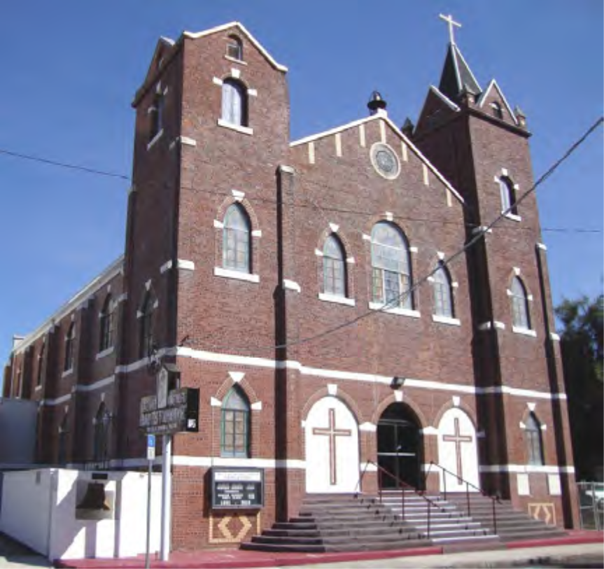 A brick church building with a tall steeple, arched windows, and cross symbols, set against a clear blue sky.