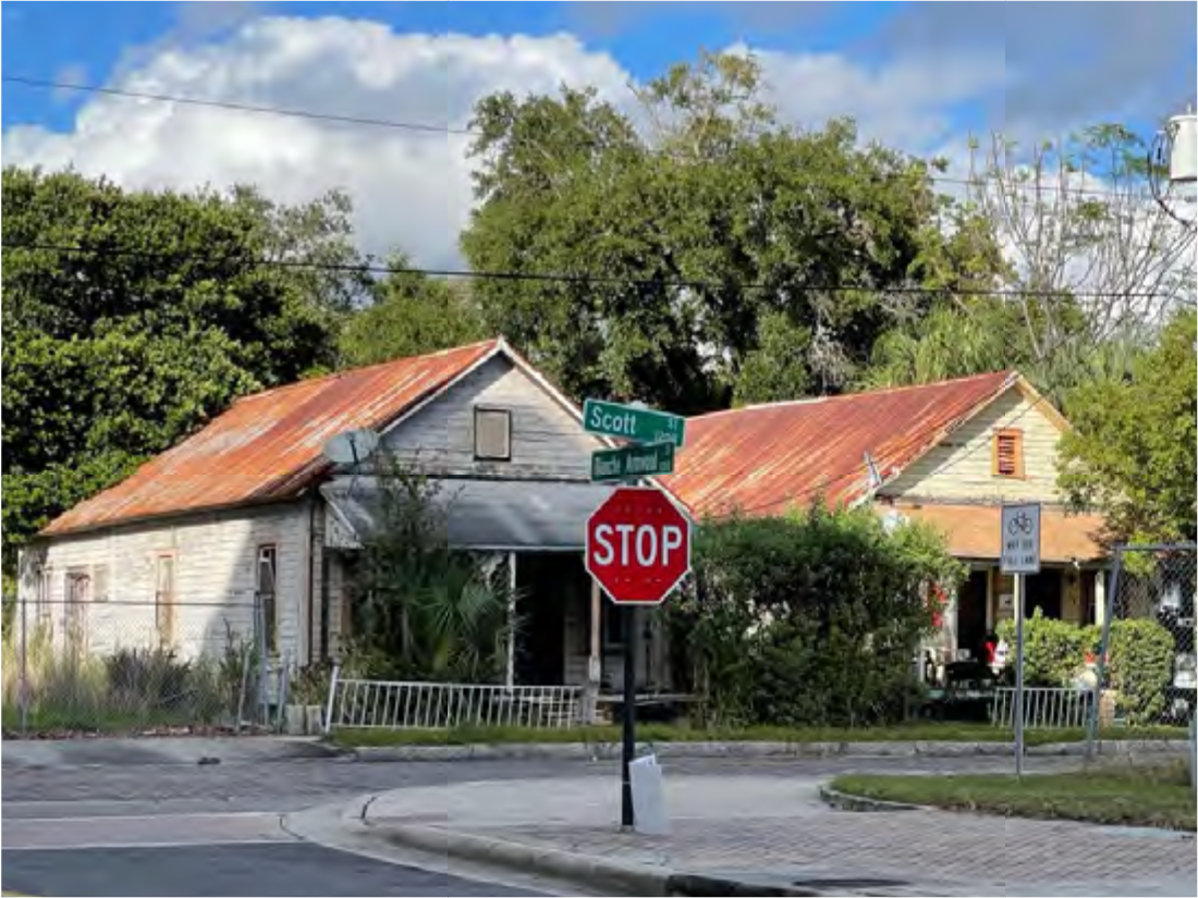 An old house with a rusted roof at a street corner with a stop sign and greenery in the background.