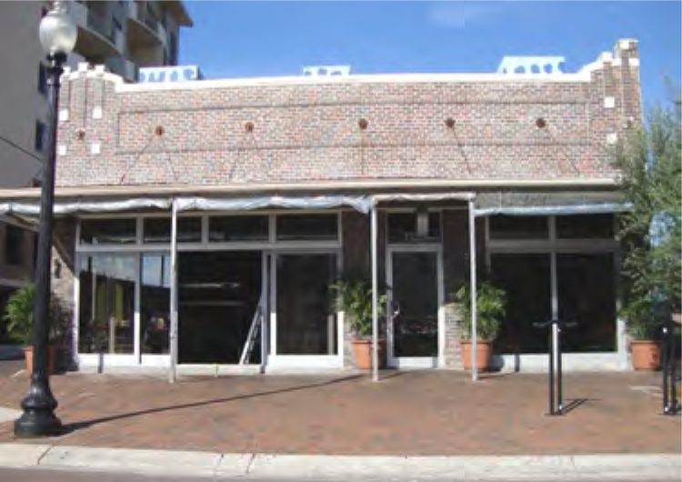 A brick building with large glass windows and a covered patio, surrounded by potted plants and street lamps.