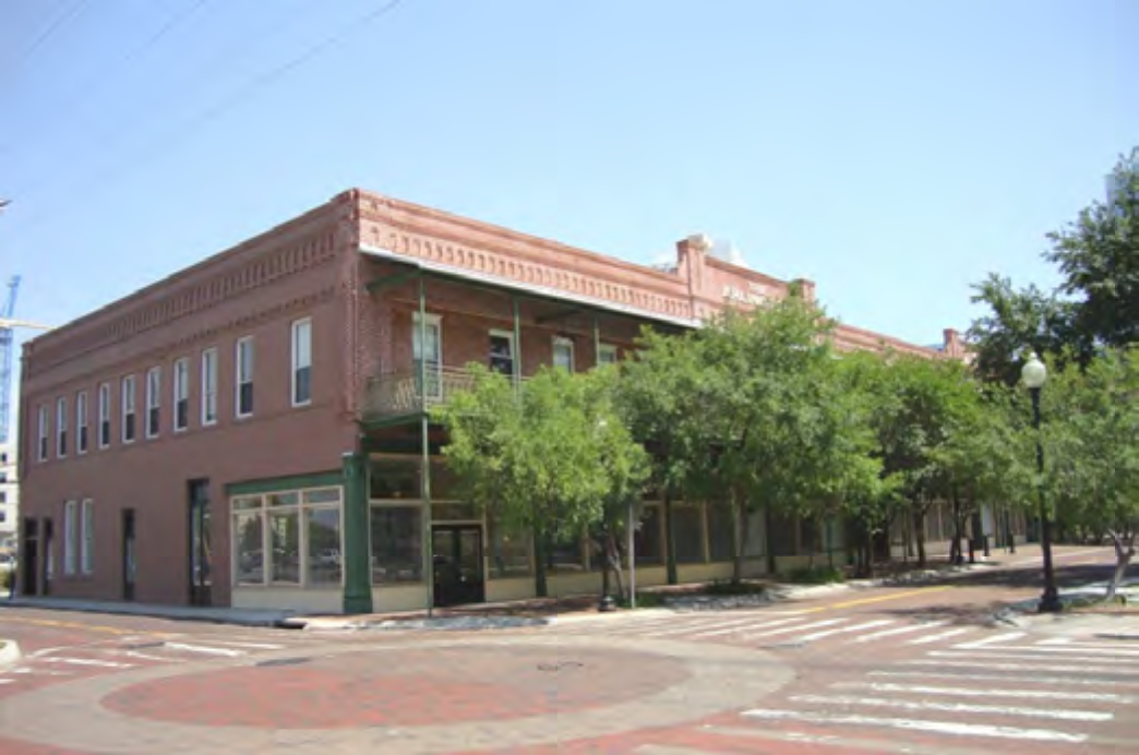 Historic brick building with green balconies and trees, located at a street intersection under a clear blue sky.