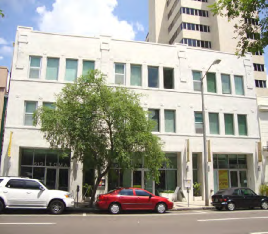 A modern white building with large windows and a tree in front, situated on a city street with parked cars.
