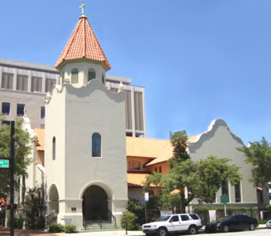 A historic church building with a tiled roof and a tall cross atop the tower, surrounded by trees and modern buildings.