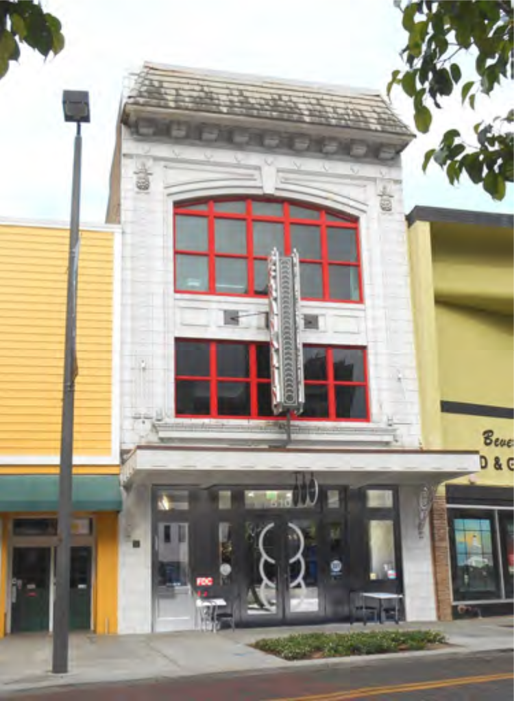 Historic building with large red windows and a marquee sign, flanked by two colorful storefronts on a street.
