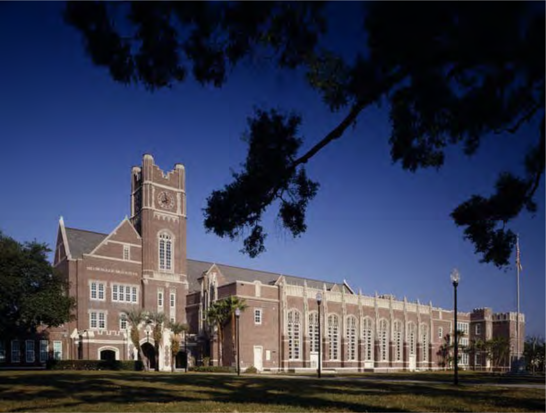 Historic brick school building with a clock tower, surrounded by greenery and blue sky.