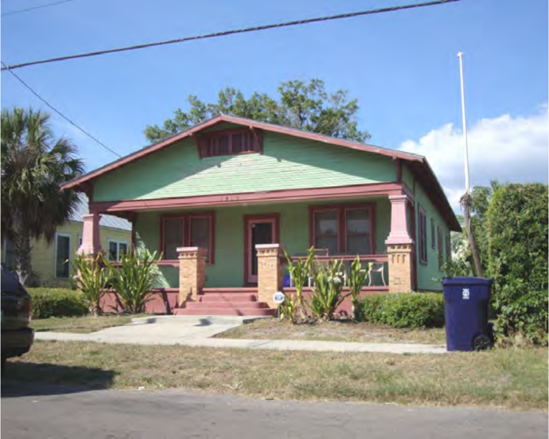 A green bungalow with pink accents, featuring a front porch and garden, under a clear blue sky.