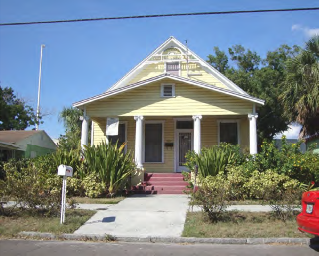 Two-story yellow house with a front porch, surrounded by greenery and a sidewalk, under a clear blue sky.