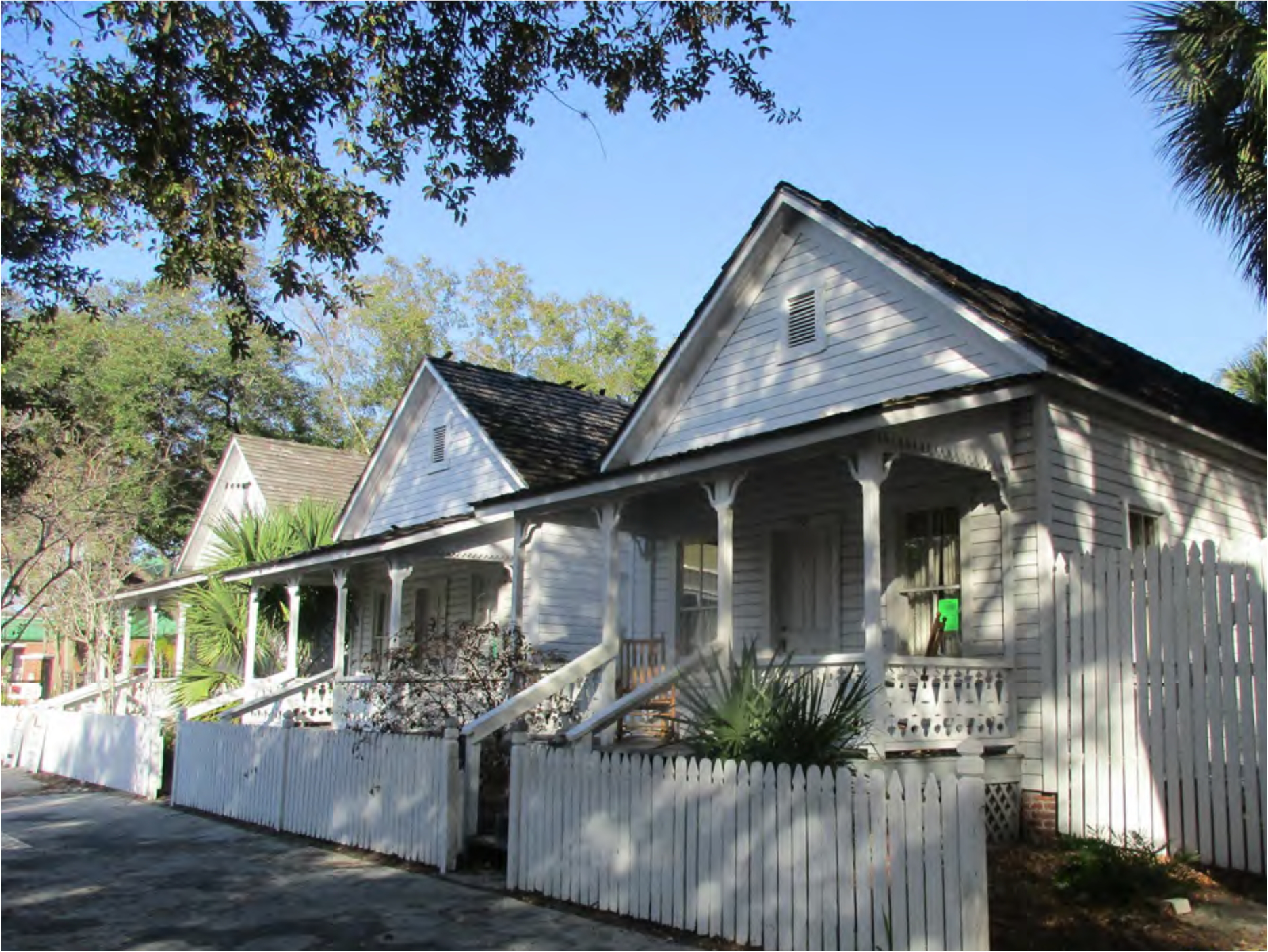 A row of white wooden houses with porches and a picket fence, surrounded by greenery under a clear blue sky.