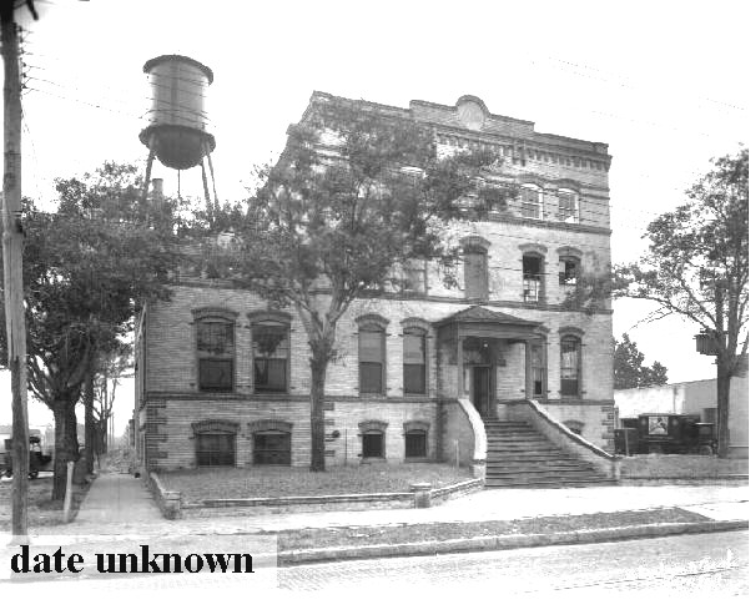 Historic brick building with a water tower, featuring a staircase entrance and trees, captured in black and white.