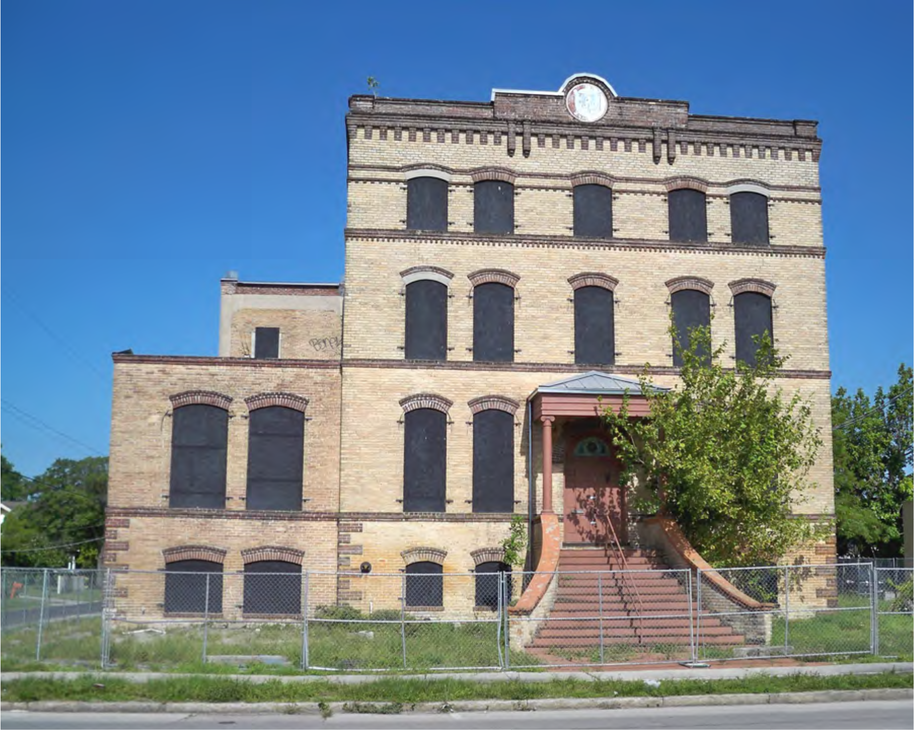 Abandoned brick building with boarded windows, a staircase, and a clock, set against a clear blue sky.