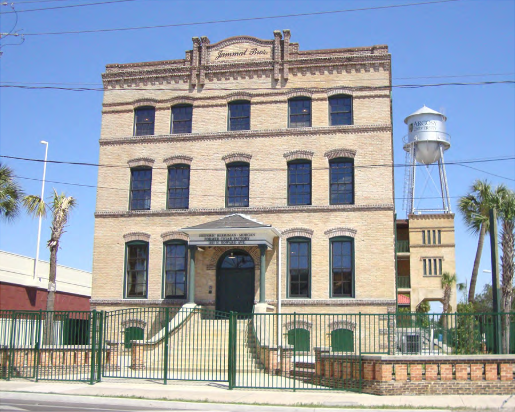 Historic brick building with arched windows and a welcoming entrance, surrounded by a green fence and a water tower in the background.