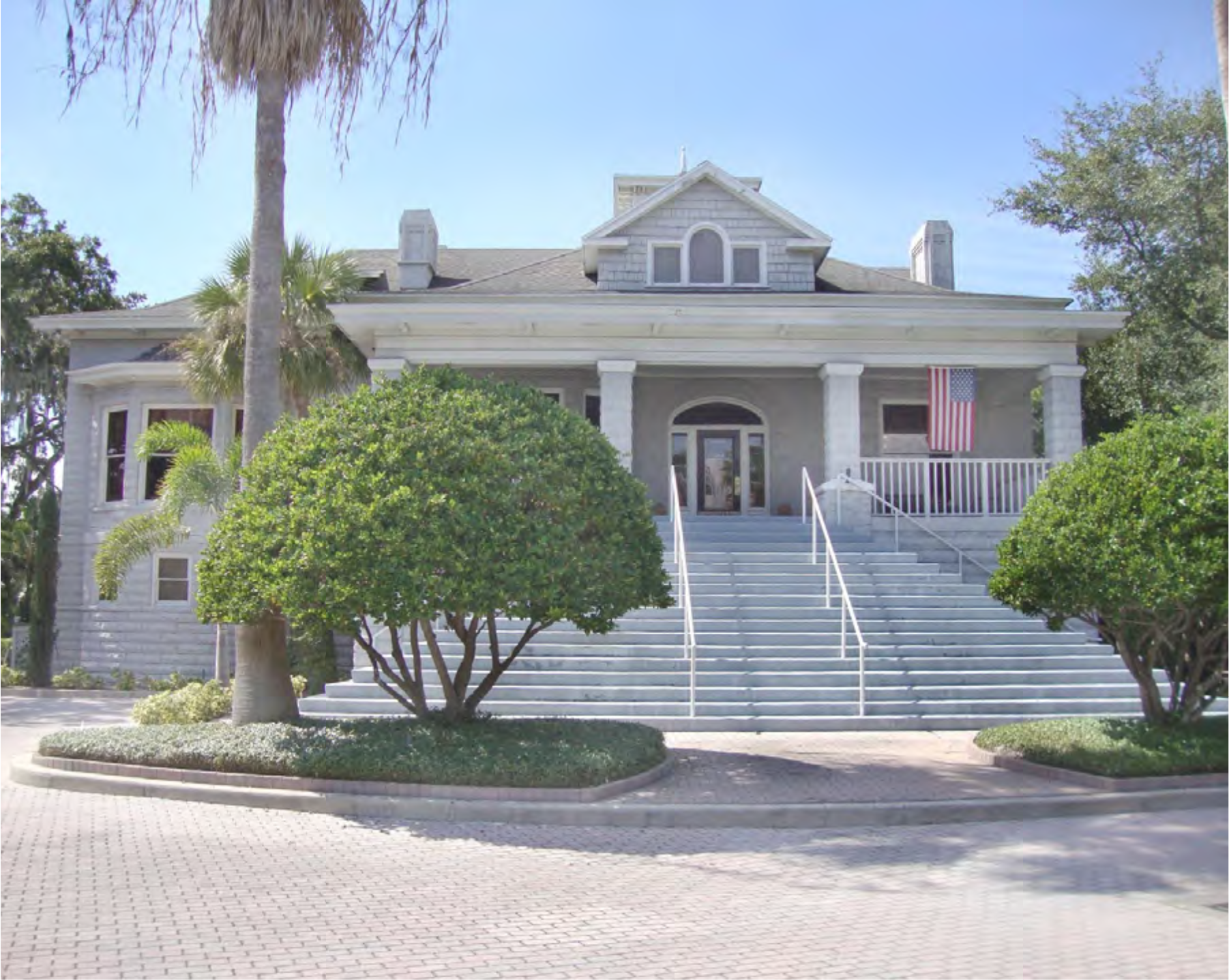 A large gray building with white columns, stairs, and a U.S. flag, surrounded by neatly trimmed bushes and palm trees.