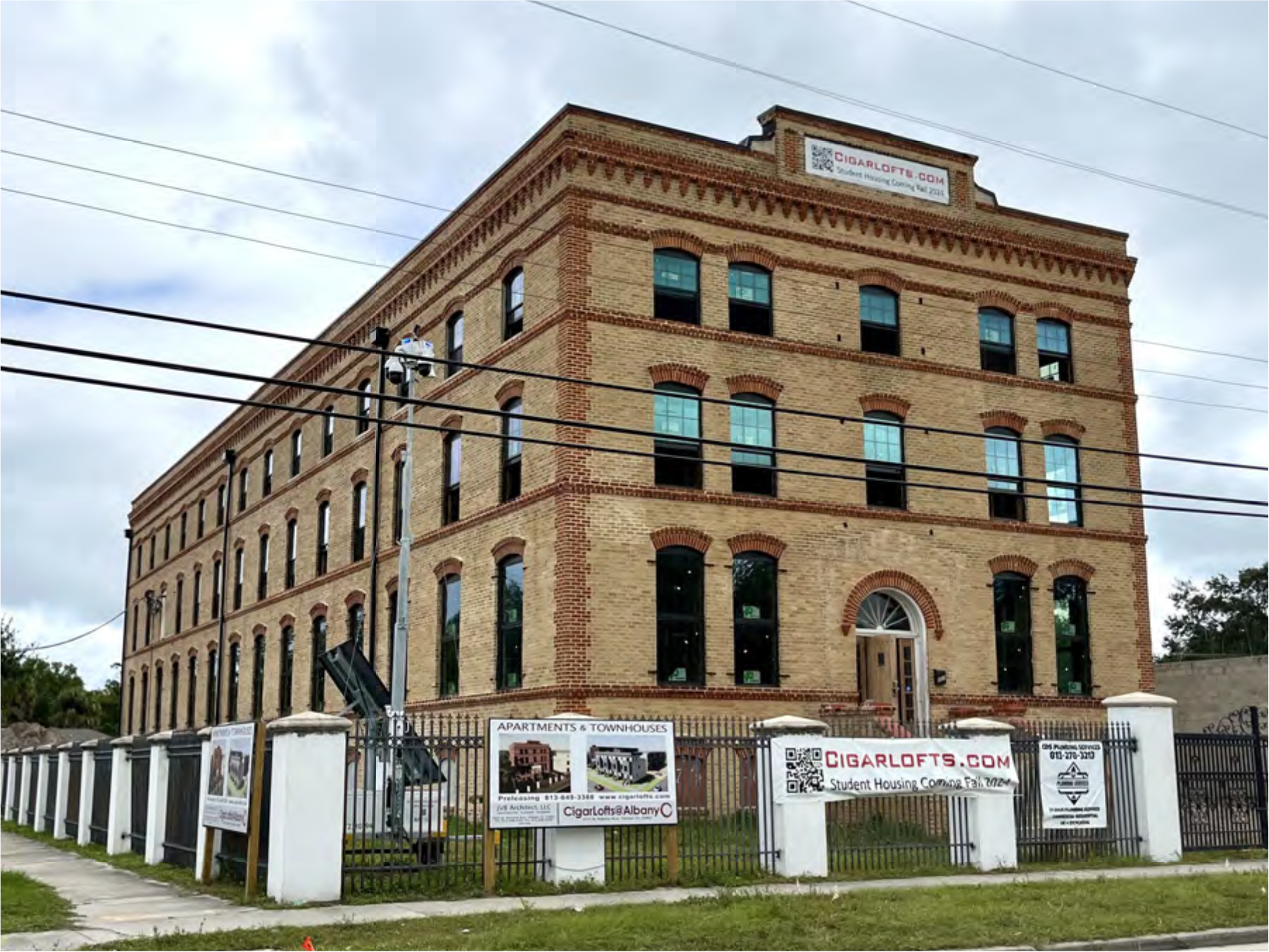 Historic brick building with large windows, featuring signs for Cigar Lofts apartments and a fenced yard.
