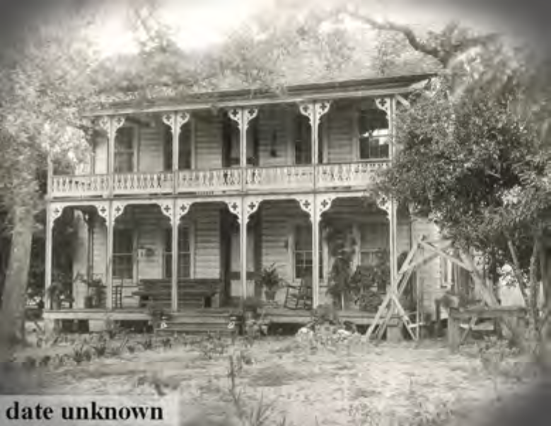 Historic two-story wooden house with a detailed porch and steps, surrounded by trees, date unknown.