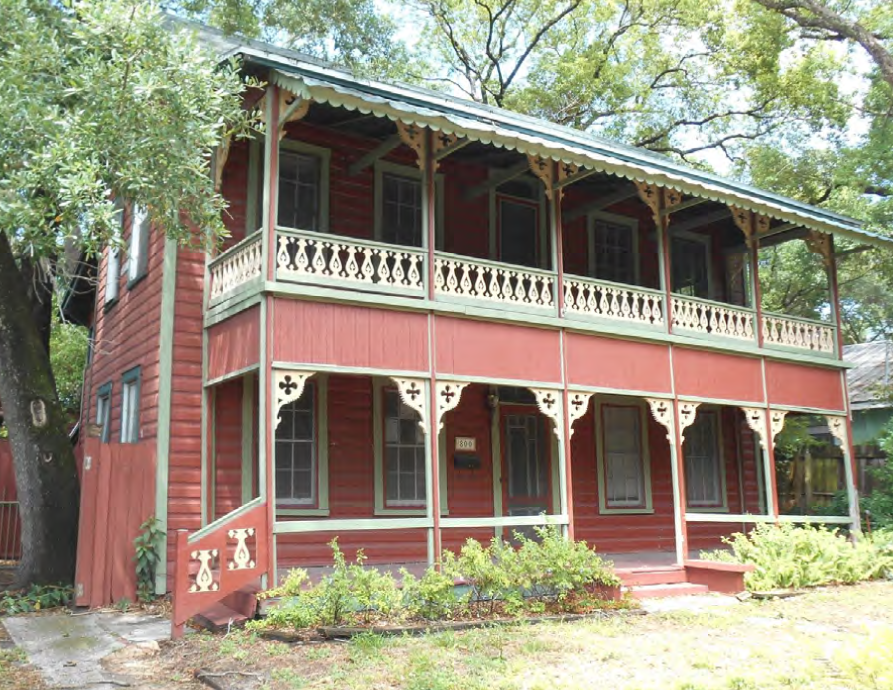 Two-story red house with white trim, featuring a large balcony, green roof, and surrounded by trees and plants.