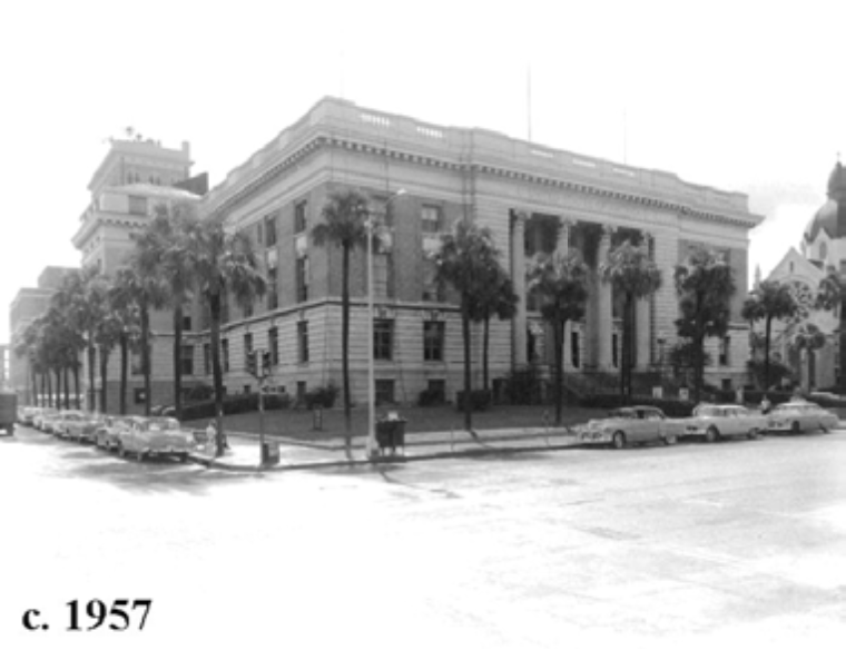 Historic building in black and white, palm trees in front, vintage cars parked on the street, circa 1957.