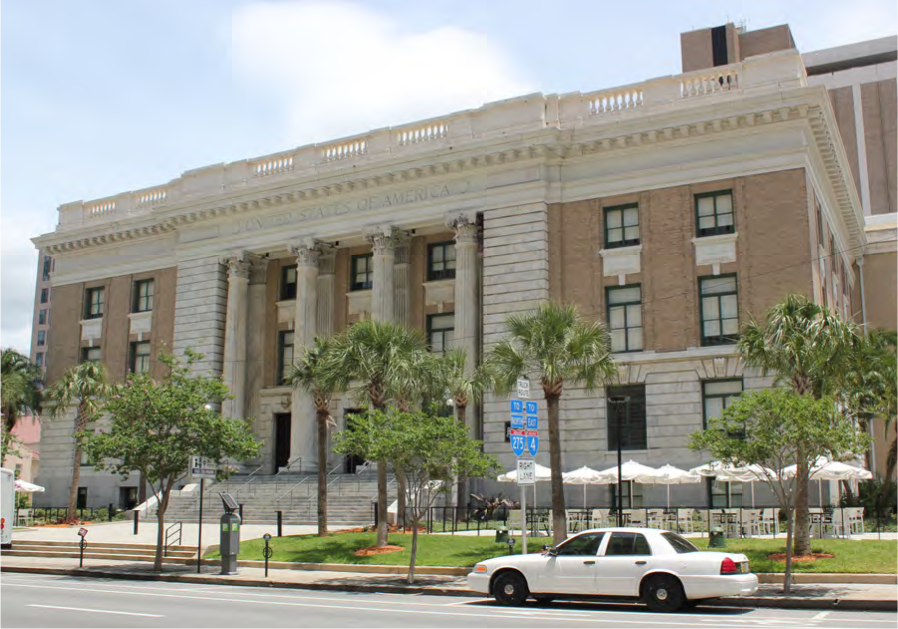 Historical building with columns and palm trees, featuring outdoor seating areas, located near a street with parked cars.