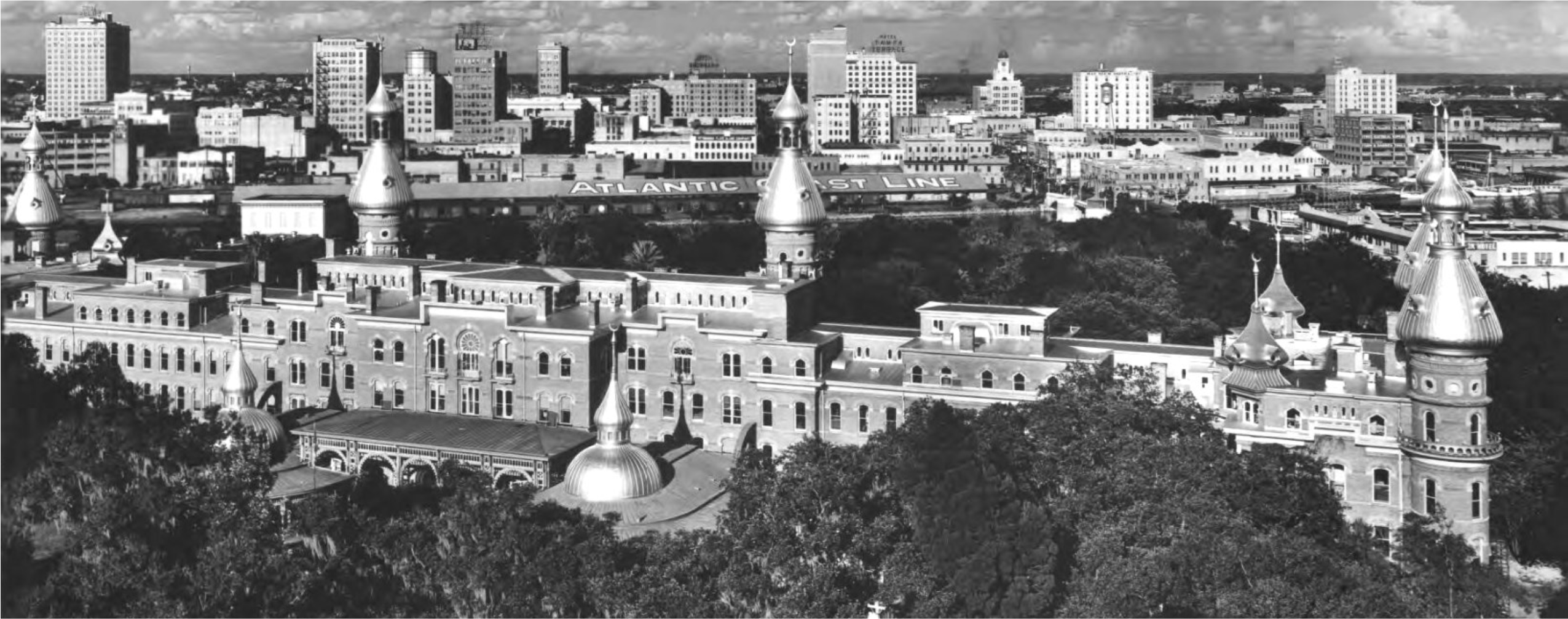 Aerial view of a historic building with towers surrounded by trees, with a city skyline and signage visible in the background.