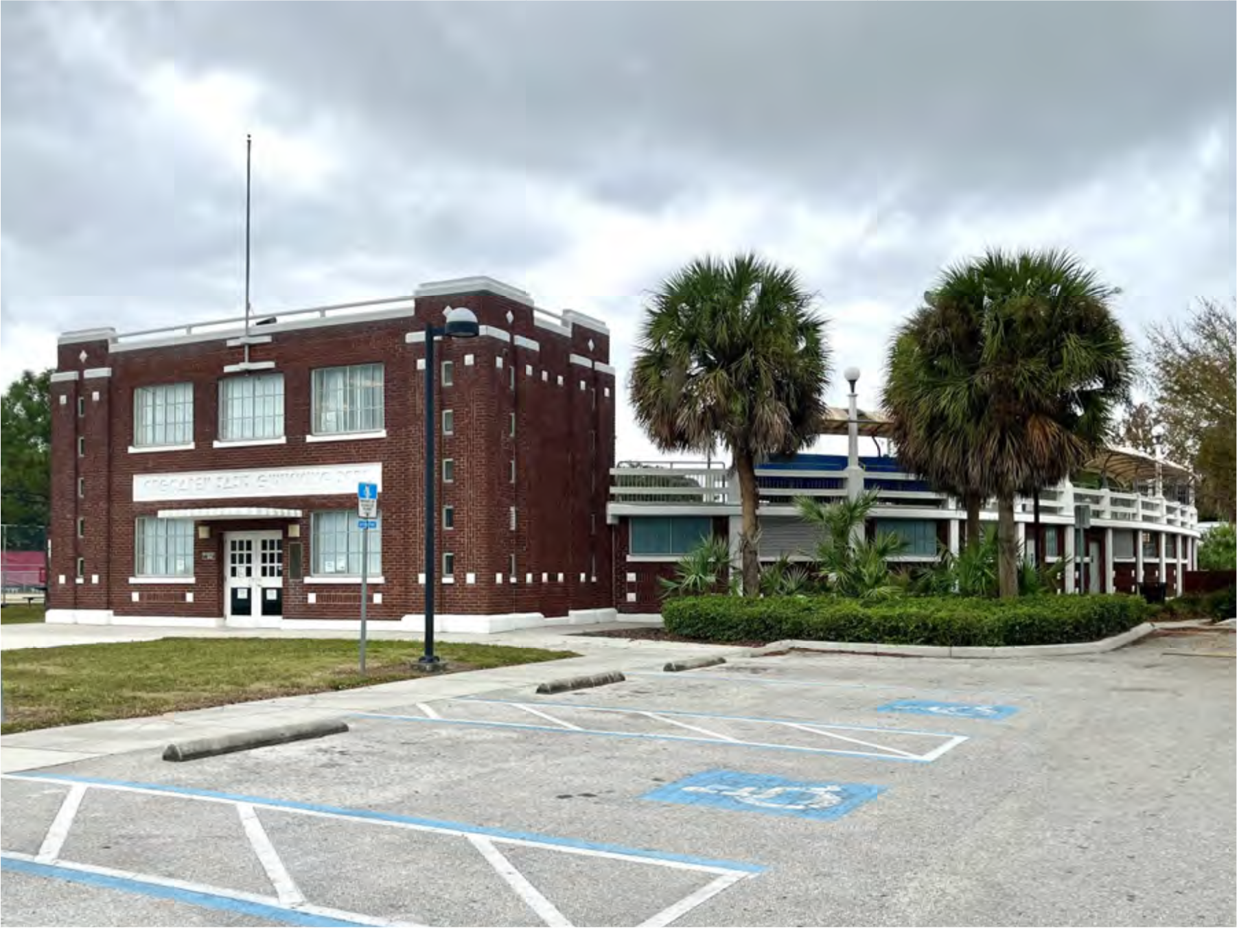 Exterior view of a brick building with palm trees, parking area, and flagpole under a cloudy sky.