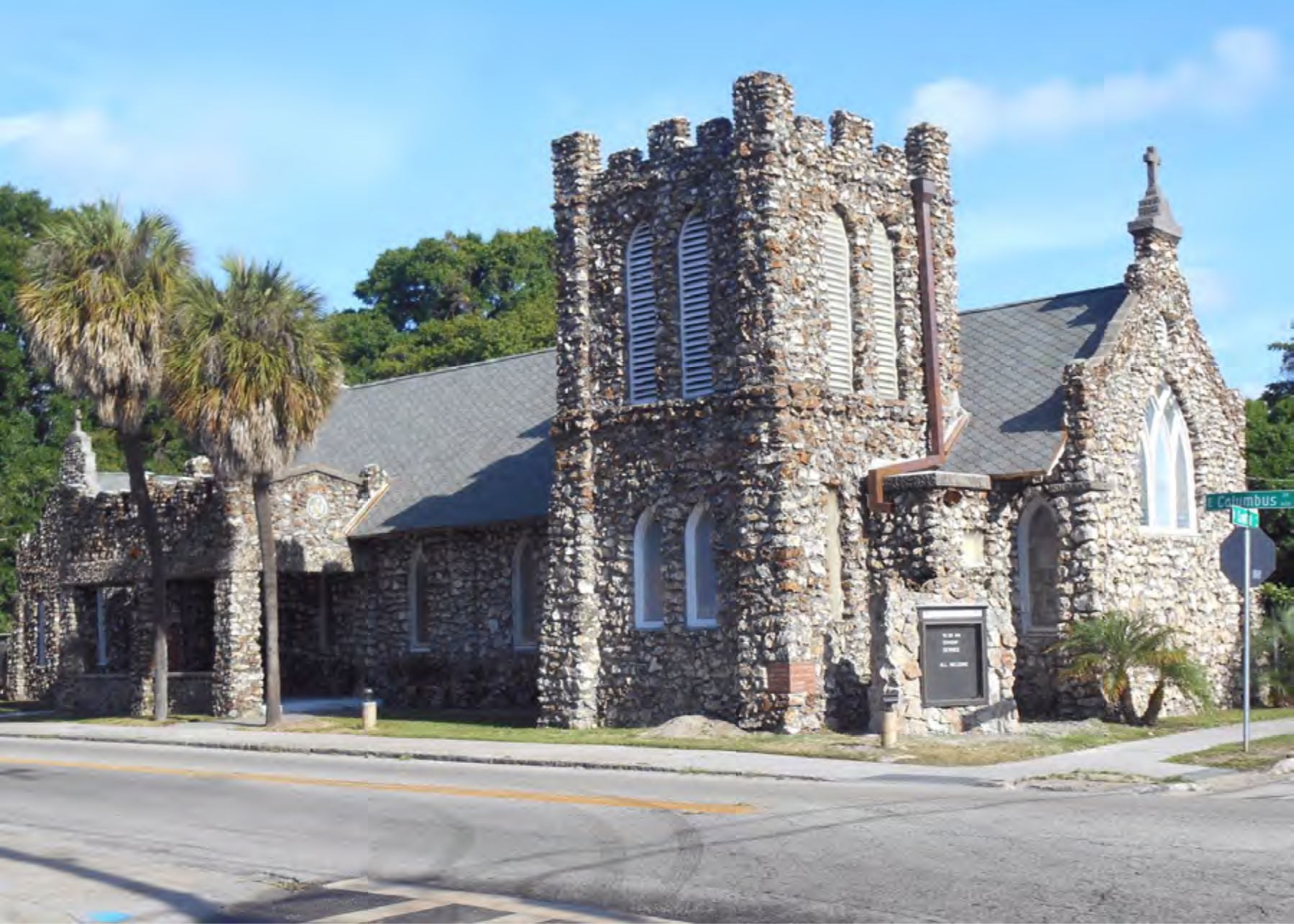 A unique stone church with a tower, surrounded by palm trees, located at a street intersection under a clear blue sky.