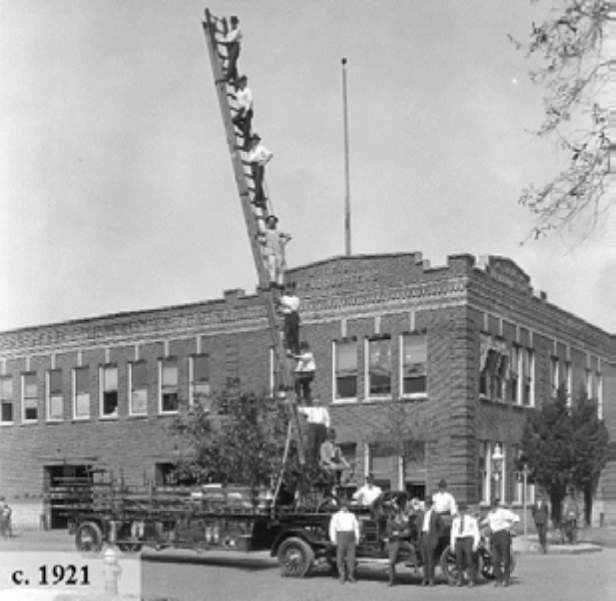 Historical photo of a large ladder truck with several men climbing the ladder, outside a brick building, circa 1921.