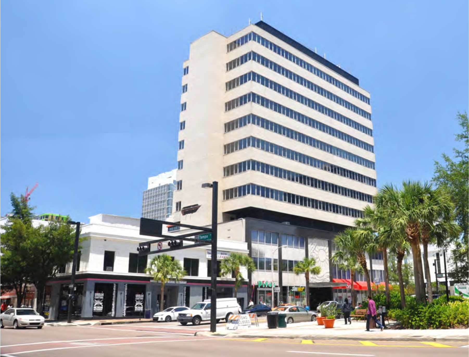 A tall modern building next to a low-rise structure and palm trees, set against a blue sky in an urban area.