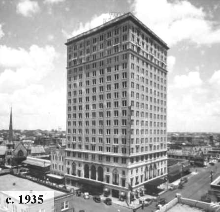 A tall historic building from 1935, surrounded by smaller structures and streets, under a partly cloudy sky.