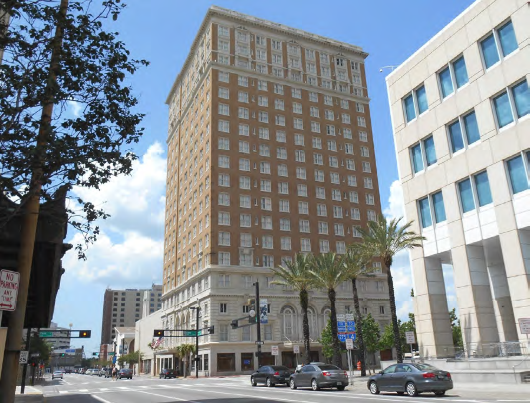 A tall, historic building with a detailed facade, surrounded by palm trees and modern architecture, under a blue sky.